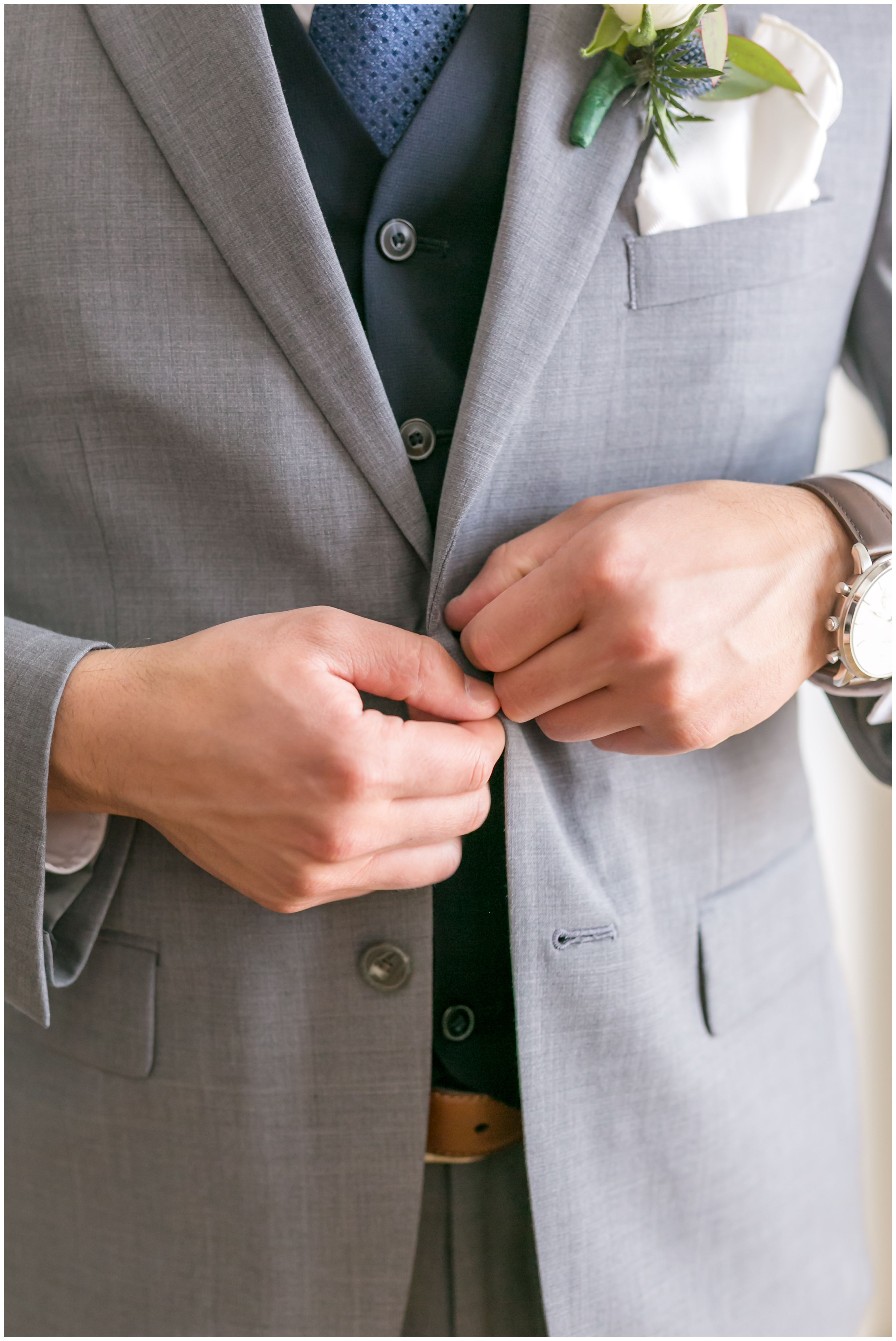 Groom getting ready buttoning light grey jacket over navy vest and tie detail